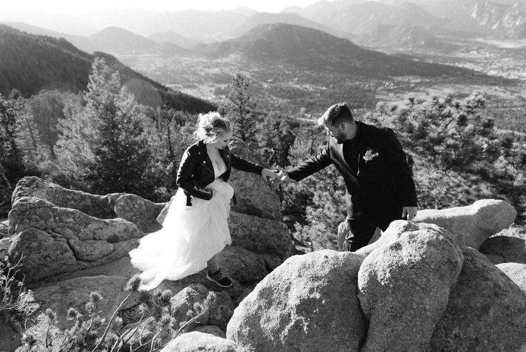Groom helps bride climb across boulders at their estes park elopement
