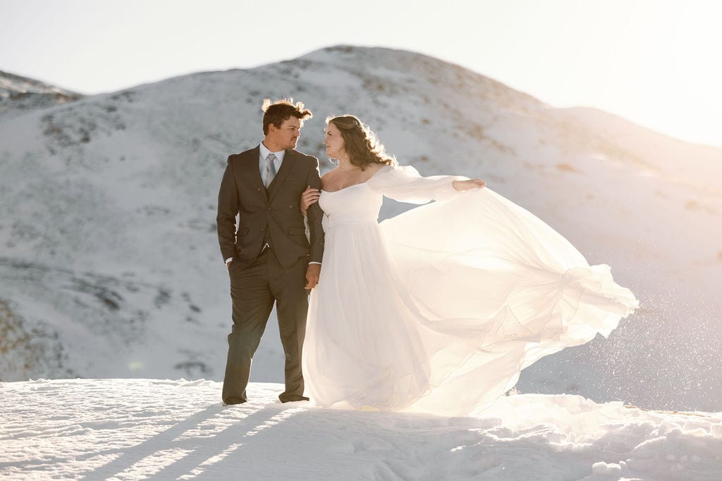 Snow covered mountain wedding portraits in the wind with dress blowing up high and couple kissing