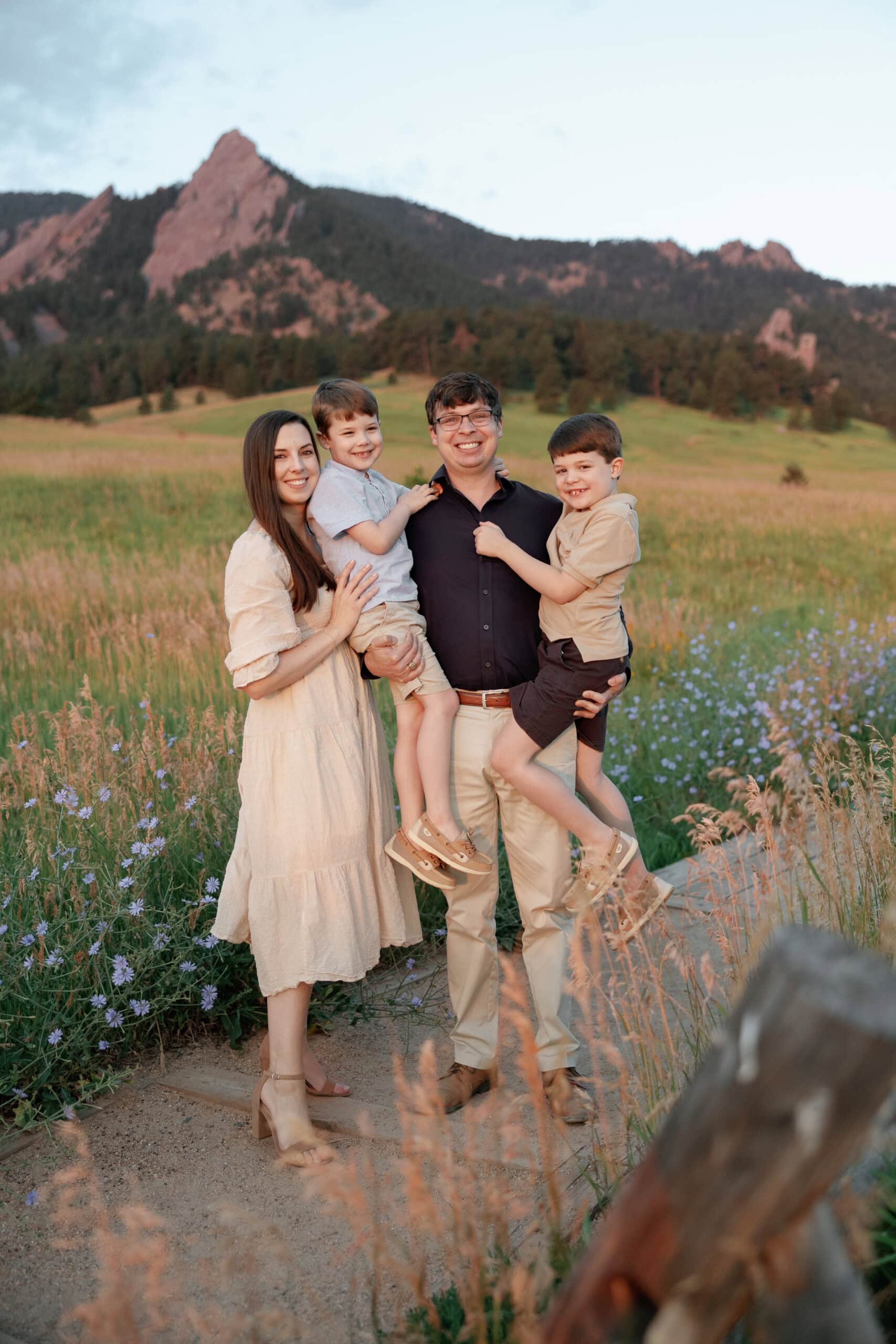 Chautauqua Park Family Photos at Sunrise in front of the flatirons. Family poses for the camera with dad holding their two boys and mom standing next to them. The sun has started to rise and the glow of the sunlight softly touches their skin