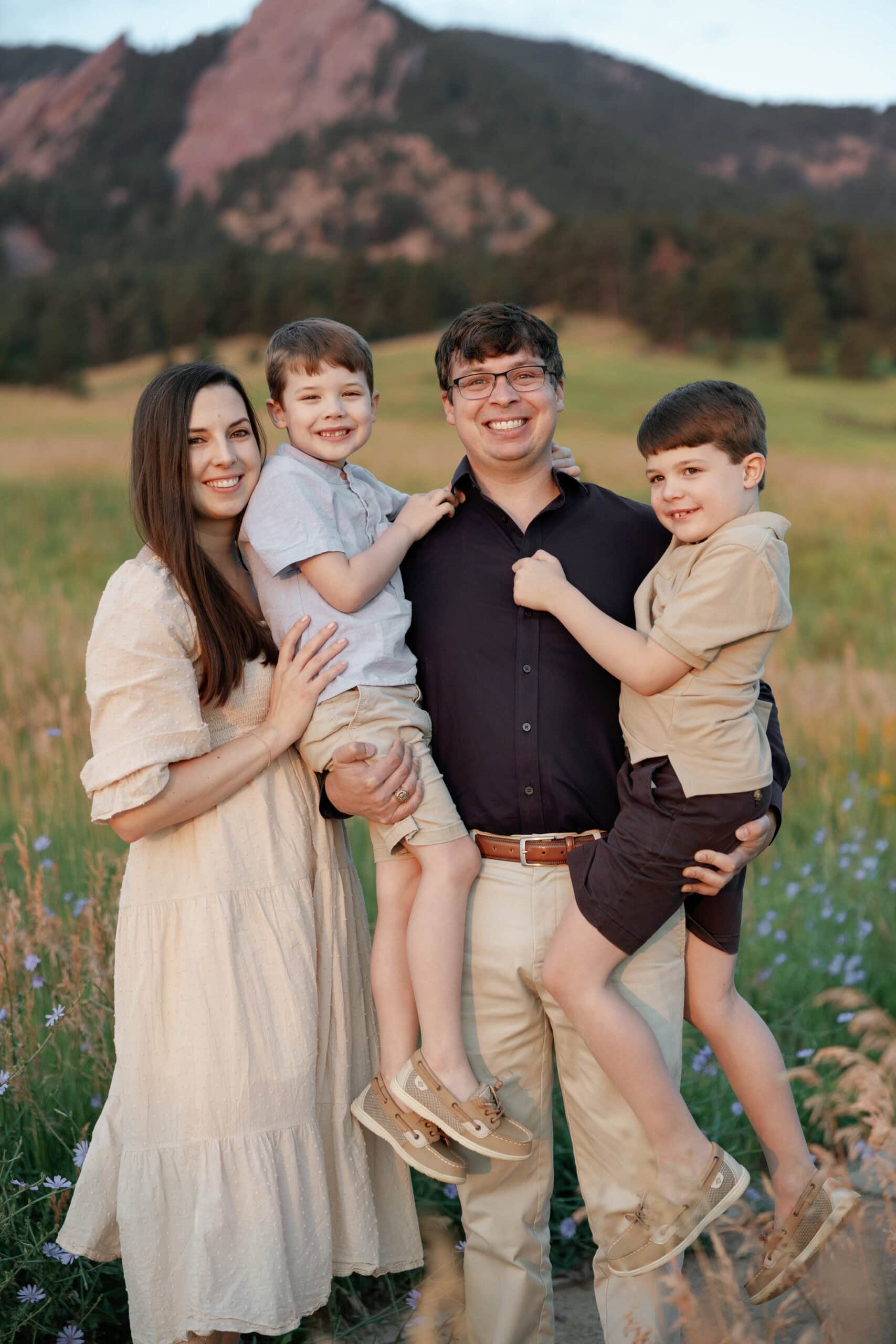 Chautauqua Park Family Photos at Sunrise in front of the flatirons. Family poses for the camera with dad holding their two boys and mom standing next to them. The sun has started to rise and the glow of the sunlight softly touches their skin