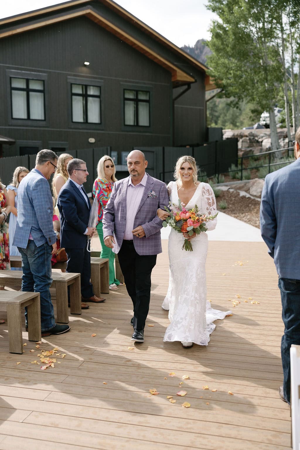 Mountain view ranch lakeview wedding ceremony in pine colorado, with country chic vibes on a cloudy day. Bride walks down the aisle with her dad carrying a colorful bouquet of flowers.