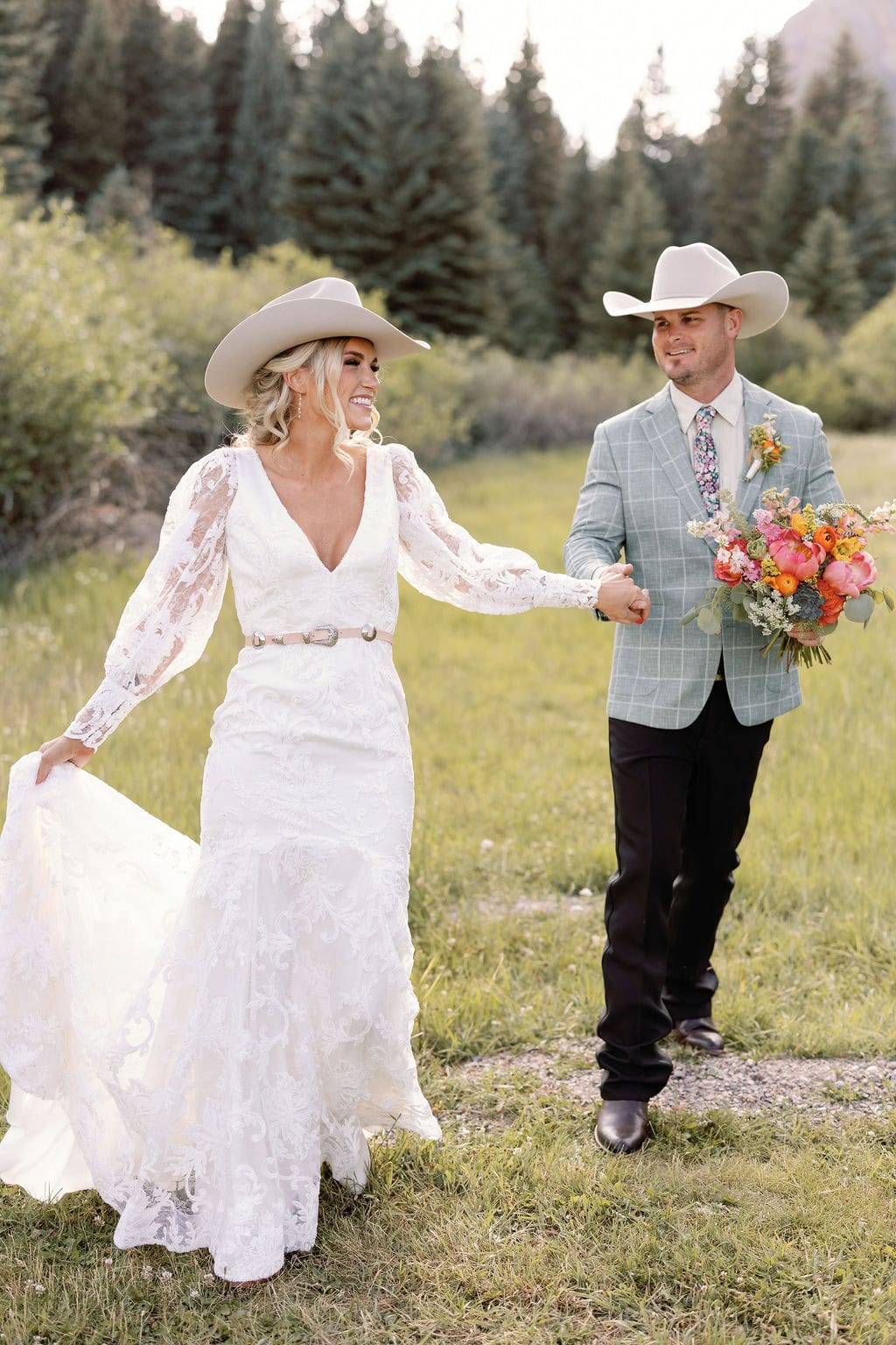 Romantic and chic country mountain wedding portraits in an open field at mountain view ranch by wedgewood weddings. Bride is with her husband as he holds colorful floral bouquet and the two are walking towards the camera laughing.