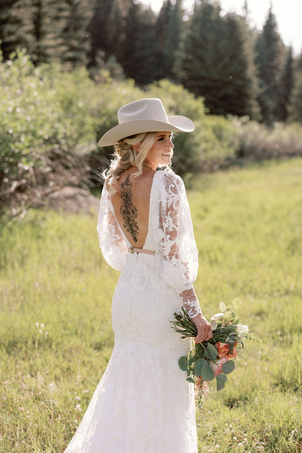 Bridal portrait with bride facing away from the camera, showcasing her gorgeous back tattoo as she looks off to the side wearing a cowboy hat. The bride is sunkissed with gorgeous sunlight in a field at her mountain view ranch wedding in pine, colorado