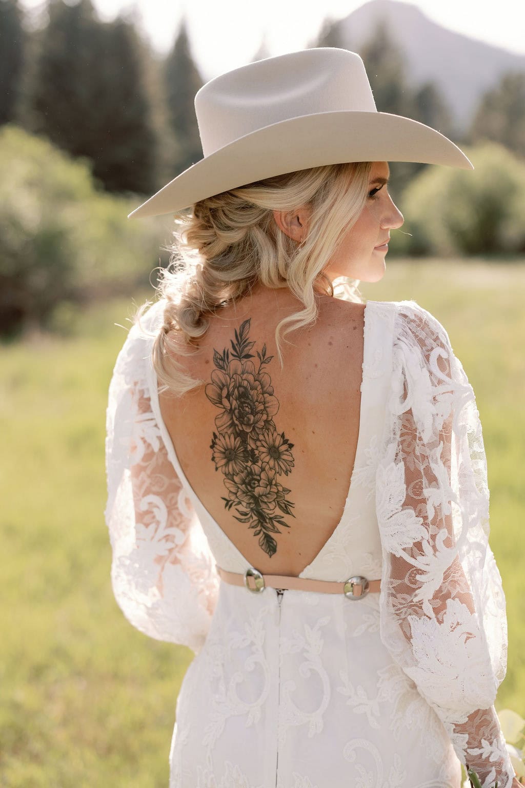 Bridal portrait with bride facing away from the camera, showcasing her gorgeous back tattoo as she looks off to the side wearing a cowboy hat. The bride is sunkissed with gorgeous sunlight in a field at her mountain view ranch wedding in pine, colorado